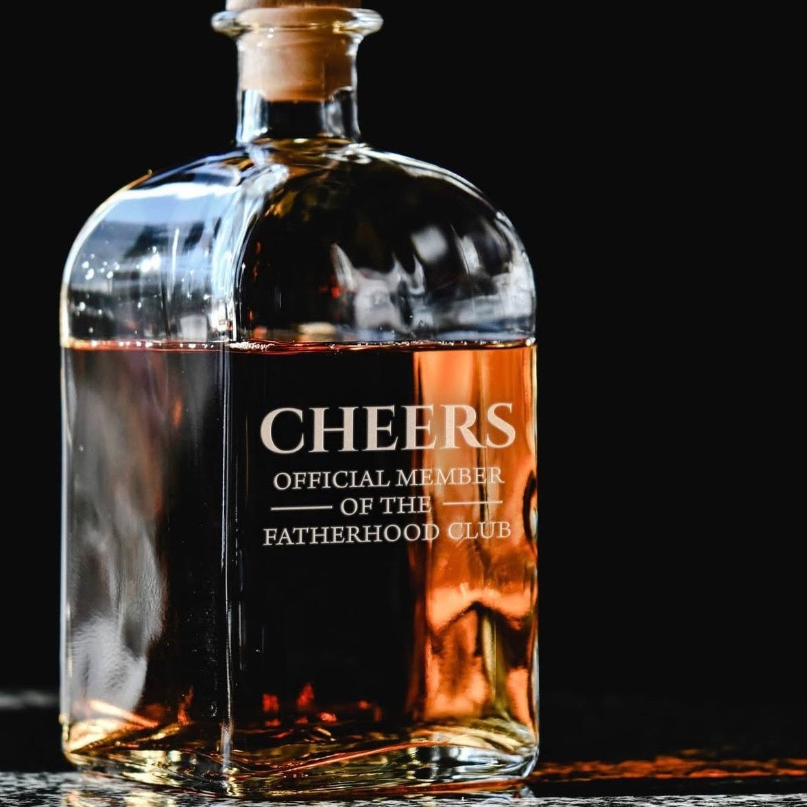 Glass decanter with brandy labeled 'Cheers Official Member of the Fatherhood Club' on a dark background