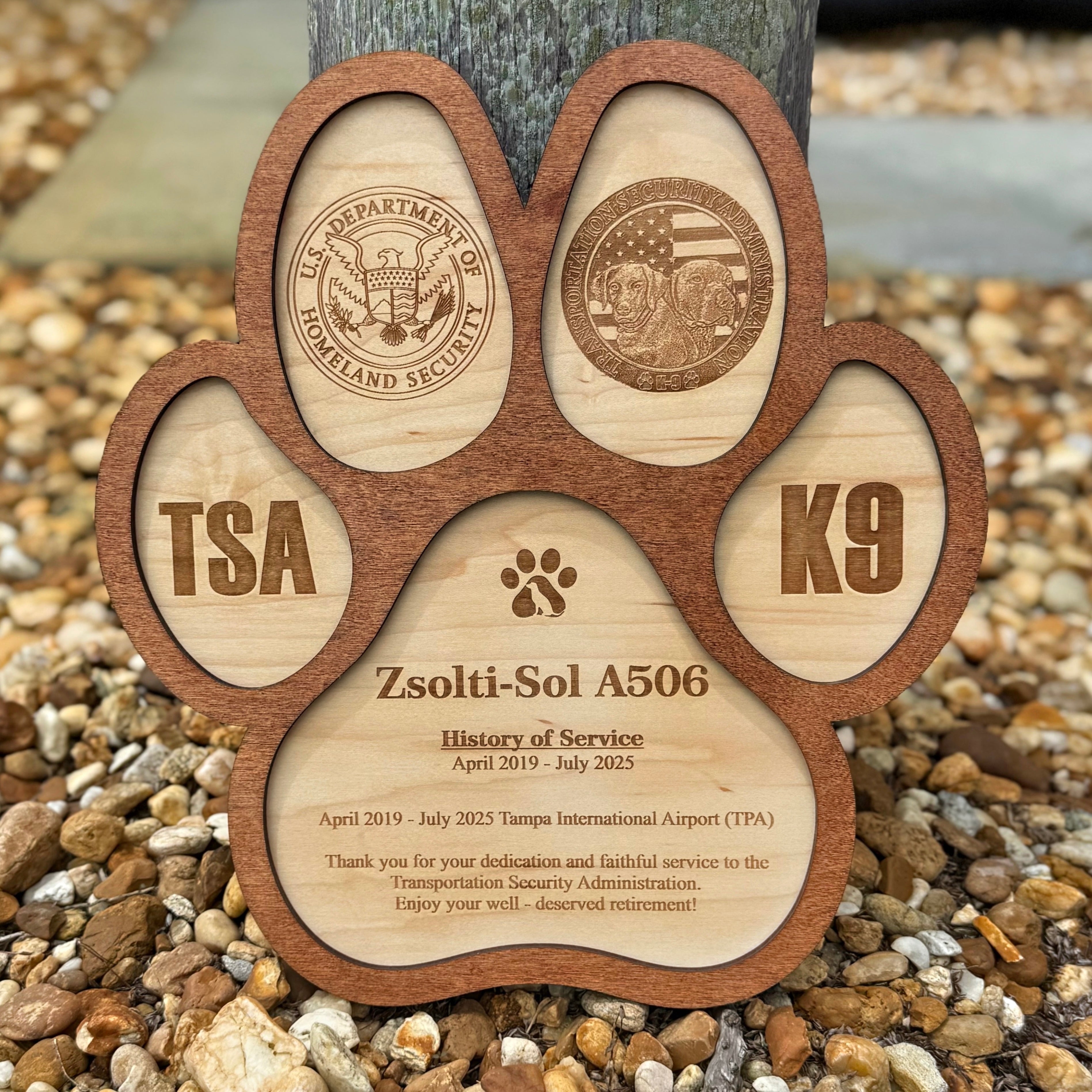 Wooden paw-shaped plaque with TSA, K9, and Department of Homeland Security logos on a pebble background.
