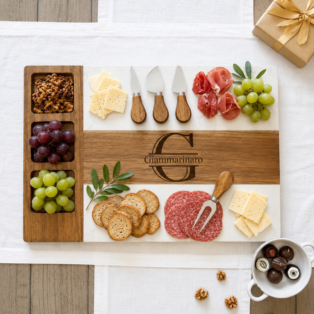 A rectangular wooden charcuterie board with a white center panel, styled with cheese, crackers, grapes, salami, prosciutto, and three cheese knives. The board is engraved with the name 'Giammarinato' and features three compartments on the left holding nuts, grapes, and grapes.